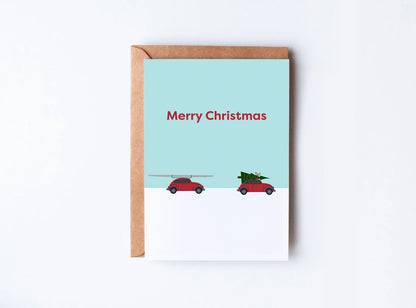 A red car with a rowing boat strapped to the top alongside a car with a christmas tree strapped to the top driving through snow. The words 'Merry Christmas' sit above.